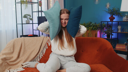 Repair work at neighbours concept. Irritated woman sit on couch cover ears with pillows annoyed by...