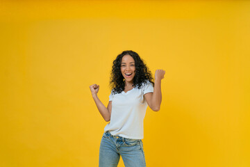 Happy curly Latin woman in white t-shirt raise hands up isolated on yellow background.