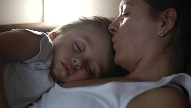Mother kissing sleeping child in forehead while napping. Parent and little boy son asleep together