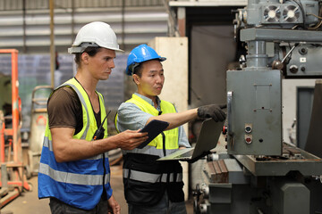 Technician engineer in protective uniform with hardhat standing and teaching apprentices or colleague worker to use computerized machine control at heavy industry manufacturing factory