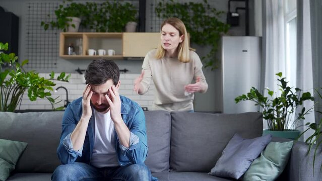 Angry young woman screams in frustration offends abases her stressed disappointed husband in the living room at home Domestic psychological violence and emotional abuse against man