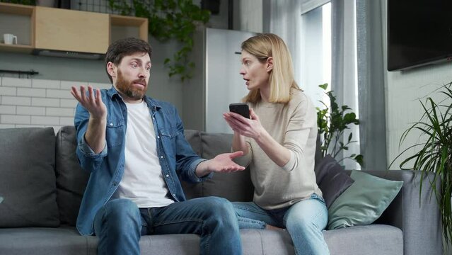 Irritated young woman finds message of infidelity on the husband phone hitting him and demands to explain Perplexed bearded man feels confused while his wife screaming at him Cheating lie concept 