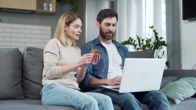 Young married couple with credit card shopping online paying bills or order food delivery using laptop sitting on the couch at home Happy family buying on internet and making payment on computer