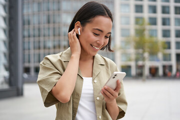 Smiling asian girl listens music in wireless headphones, looks at her phone, choosing music or...