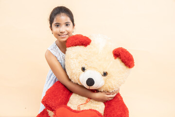 Happy indian girl kid hugging her big teddy bear isolated over beige studio background.