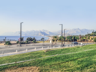 Promenade of Konyaalti Plaji overlooking the mountains and the beach in Antalya, Turkey. The car drives along the city road against the backdrop of the Mediterranean Sea and the park. Lawn with grass
