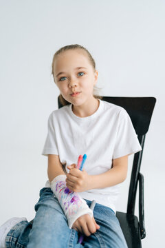 Vertical portrait of positive little girl drawing cute image with colorful marker on broken arm wrapped in white plaster bandage looking at camera. Concept of child insurance and healthcare.