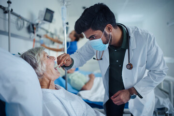 Young doctor examining throat to senior patient in hospital room.