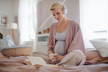 Pregnant woman sitting in her bed, listening music and reading book, enjoying time for herself.