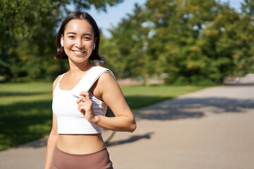 Smiling asian fitness girl holding towel on shoulder, workout in park, sweating after training...