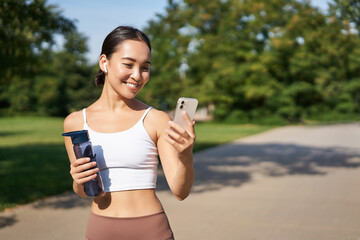 Fitness woman with water bottle and smartphone, jogging in park and smiling, looking at her mobile...