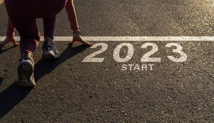 Word 2023 written on the asphalt road and athlete woman runner preparing to start on an athletics track engraved for new year at sunset.Concept of new year 2023,challenge or career path and change.