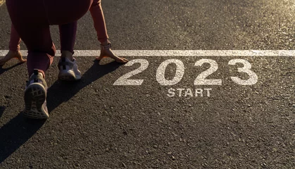 Tuinposter Word 2023 written on the asphalt road and athlete woman runner preparing to start on an athletics track engraved for new year at sunset.Concept of new year 2023,challenge or career path and change. © Yingyaipumi