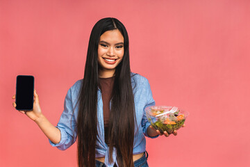 Clean eating diet concept. Asian woman holding vegeterian salad or bowl in take away container. Close up, copy space, isolated over pink background. Showing mobile phone screen.