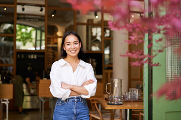 Portrait of young businesswoman in her own cafe, manager standing near entrance and inviting you,...