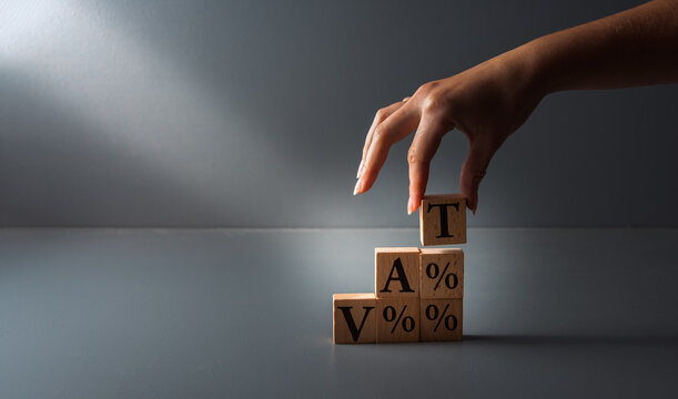 hand posing wooden blocks with vat or value added tax . concept of paying taxes , on grey background .