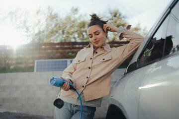 Young woman holding power supply cable from her electric car, prepared for charging it in home,...