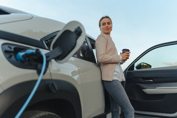 Young woman with cup of coffee waiting while her electric car charging, sustainable and economic...