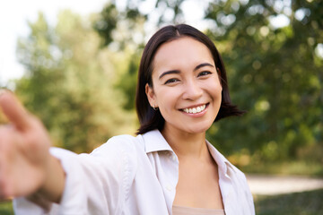 People and lifestyle. Happy asian woman takes selfie in park, photo on smartphone, smiling and looking joyful