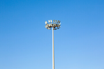 An electric pole with many spotlights on it during the daytime against a blue sky.