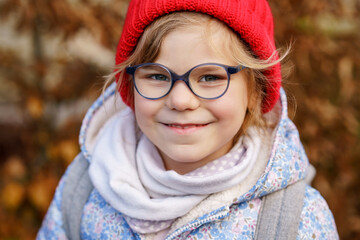 Portrait of a cute preschool girl with eye glasses and red hat outdoors. Happy funny child wearing new blue glasses. Autumn day in the city.