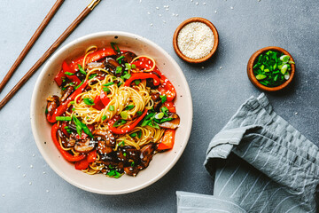 Vegetarian stir fry noodles with vegetables, paprika, mushrooms, chives and sesame seeds in bowl. Gray table background, top view, copy space