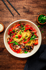 Vegan Stir fry noodles with vegetables, paprika, mushrooms, chives and sesame seeds in bowl. Wooden table background, top view, copy space
