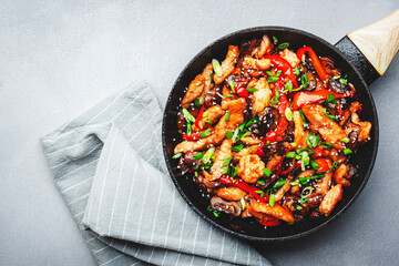 Stir fry with chicken, red paprika, mushrooms and chives in frying pan. Asian cuisine dish. Gray kitchen table background, top view, copy space