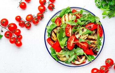 Healthy vegan salad with grilled vegetables, paprika, zucchini, eggplant with fresh tomatoes and mixed herbs. White table background, top view