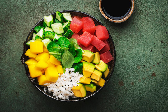 Trendy hawaiian poke bowl with tuna, avocado, mango, cucumber, lamb lettuce and white rice. Soy sauce and sesame dressing. Old green table background, top view
