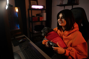 gamer girl in orange sweatshirt plays video games on her computer in her set-up with her pet on her lap.
