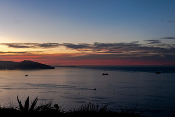 Fototapeta premium Twilight sunset colorful cloudy sky with boats floating and bay of water with a mountain shore. Blue sky, red and orange colors. Akid Lotfi public park in Oran, Algeria. Herbs in backlit foreground.