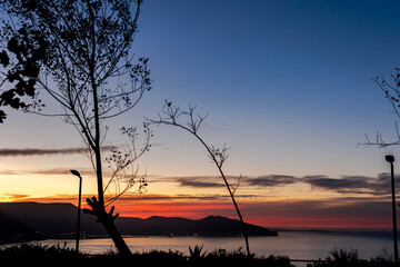 Twilight sunset colorful cloudy sky with boats floating and bay of water with a mountain shore. Blue sky, red and orange colors. Akid Lotfi public park in Oran, Algeria. Trees in backlit foreground.