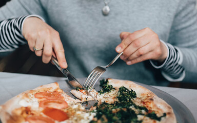 Female person cutting delicious pizza with tomato and spinach. Close-up of hands using fork and knife.
