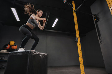 Fototapeta na wymiar Woman doing box jump exercise as part of her crossfit training. Female athlete doing squats and jumping onto the box in dark workout gym. Copy space