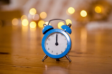 Fototapeta na wymiar Blue arrow alarm clock on wooden floor, background of Christmas tree, bed, garlands, yellow bokeh, midnight, new year