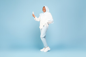 Happy african american woman in hijab using cellphone, standing on blue studio background, free space, full body length