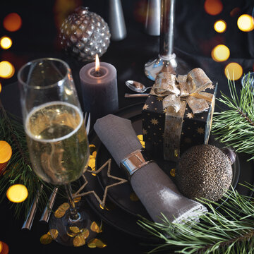 Atmospheric table decoration for a christmas or new year dinner. Place setting in modern style. Champagne, gift box, festive branch decoration with candle and golden bokeh on black tablecloth.