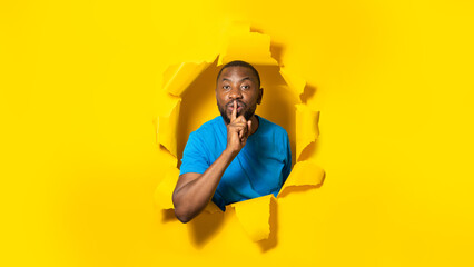 Black man making silence gesture telling secret, breaking through hole of yellow paper sheet,...