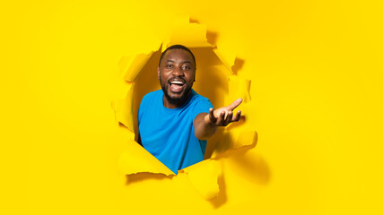 Happy black man stretching open palm, holding invisible object, posing through hole in torn yellow...