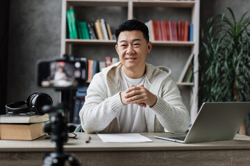 Handsome asian man in casual clothes talking and gesturing while recording video on modern phone. Male blogger sitting at home and doing live stream.