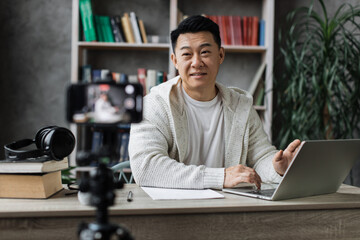 Attractive asian male in casual clothes sitting at desk working on laptop and recording video on digital smartphone. Concept of freelance and modern technology.