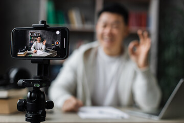 Smiling asian man in casual clothes sitting at desk with books, notes and laptop recording video blog and showing sign ok. Female tutor working at home during distance learning.