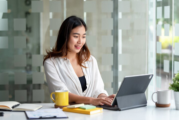 Attractive Asian business woman sitting at work happily working using tablet at office and smiling comfortably.
