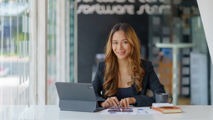 Attractive Asian business woman sitting at work happily working using tablet at office and smiling comfortably.