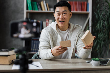 Handsome asian man in casual wear recording video on camera while unpacking gift box. Male blogger sharing his emotions with his subscribers in social networks.