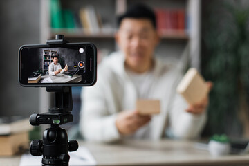 Focus on smart phone screen happy asian man in casual wear recording video on camera while unpacking gift box. Male blogger sharing his emotions with hs subscribers in social networks.