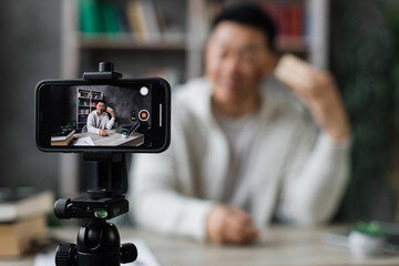 Focus on smart phone screen happy asian man in casual wear recording video on camera while...