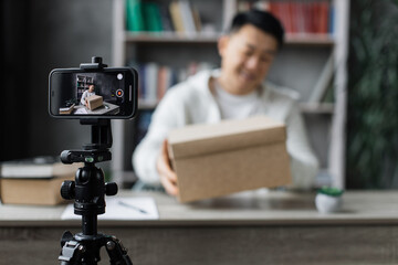 Focus on smart phone screen happy asian man in casual wear recording video on camera while unpacking gift box. Male blogger sharing his emotions with hs subscribers in social networks.