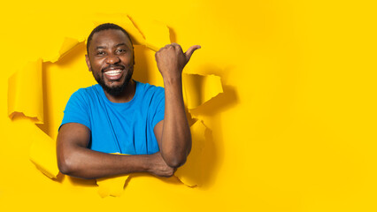 Excited african american man posing through hole in torn yellow paper, pointing thumb at free...